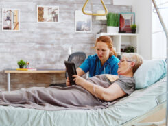 Eine Krankenschwester hilft einem älteren Patienten in einem Krankenhausbett bei der Bedienung eines Tablets. Der Patient trägt eine Brille und Kopfhörer, während die Krankenschwester auf den Bildschirm zeigt. Der Raum ist hell und gemütlich, im Hintergrund sind Bilder, Pflanzen und Regale zu sehen. -huggii