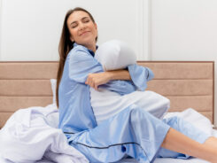 Eine Frau in hellblauem Schlafanzug sitzt auf einem Bett, umarmt ein weißes Kissen und lächelt zufrieden, umgeben von weißer Bettwäsche in einem hellen, modernen Schlafzimmer. -huggii