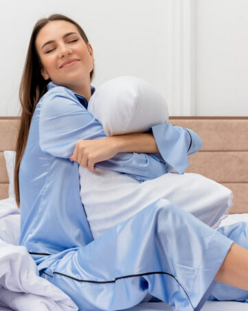 Eine Frau in hellblauem Schlafanzug sitzt auf einem Bett, umarmt ein weißes Kissen und lächelt zufrieden, umgeben von weißer Bettwäsche in einem hellen, modernen Schlafzimmer. -huggii