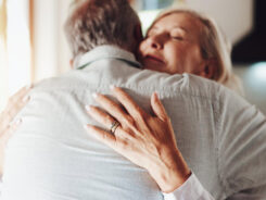 Eine ältere Frau und ein älterer Mann umarmen sich herzlich in einem sonnendurchfluteten Raum. Die Frau hat die Augen geschlossen und lächelt sanft, während ihre Hand auf dem Rücken des Mannes ruht. -huggii