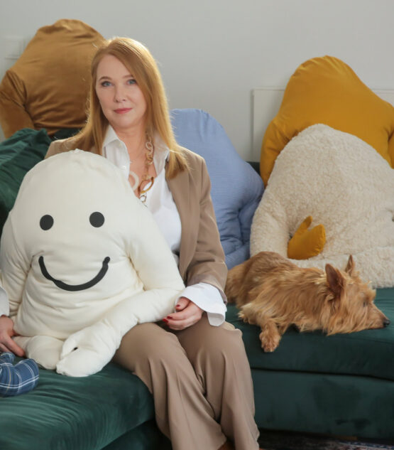 Eine Frau in einem beigen Kostüm sitzt auf einem grünen Sofa und hält ein großes, lächelndes Plüschtier im Arm. Neben ihr liegt ein Hund, und in dem gemütlichen Wohnzimmer stehen weitere Plüschkissen sowie ein Beistelltisch mit Blumen. -huggii