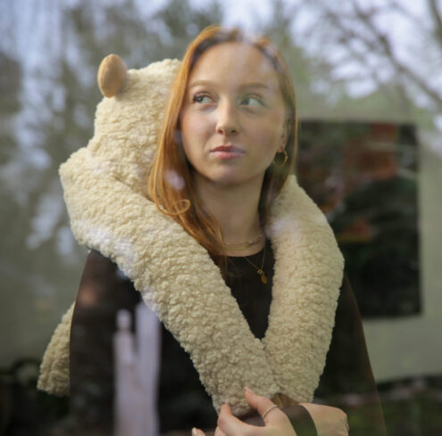 Eine Frau mit roten Haaren, die einen flauschigen, cremefarbenen Schal trägt, steht drinnen und blickt nachdenklich aus dem Fenster. Auf der Scheibe spiegeln sich Bäume und eine verschwommene Außenansicht wider. -huggii