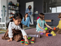 Drei Kinder spielen auf einem Teppichboden mit buntem Holzspielzeug, während zwei Erwachsene im Hintergrund in einem Klassenzimmer oder einer Kindertagesstätte zusehen. Hinter ihnen sind Regale mit Büchern und Spielzeug zu sehen. -huggii