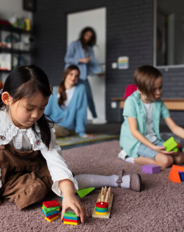 Drei Kinder spielen auf einem Teppichboden mit buntem Holzspielzeug, während zwei Erwachsene im Hintergrund in einem Klassenzimmer oder einer Kindertagesstätte zusehen. Hinter ihnen sind Regale mit Büchern und Spielzeug zu sehen. -huggii
