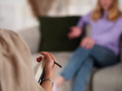 Eine Person hält ein Notizbuch und einen Bleistift in der Hand und macht sich Notizen, während eine andere Person im Hintergrund auf einem Sofa sitzt und während eines Gesprächs oder einer Therapiesitzung mit einer Hand gestikuliert. -huggii