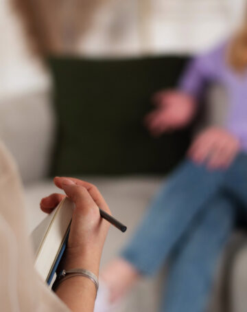 Eine Person hält ein Notizbuch und einen Bleistift in der Hand und macht sich Notizen, während eine andere Person im Hintergrund auf einem Sofa sitzt und während eines Gesprächs oder einer Therapiesitzung mit einer Hand gestikuliert. -huggii